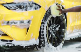 Hands of African man holding yellow sponge, washing car wheel with foam. Cleaning of modern rims of luxury yellow car at self car wash service outdoors.